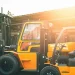 Front-right view of an orange forklift with a protective cab in an outdoor industrial yard, sun glare on the right.