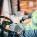 Worker in a bright safety vest and gloves grips a steering wheel inside a vehicle cab, ready to operate equipment.