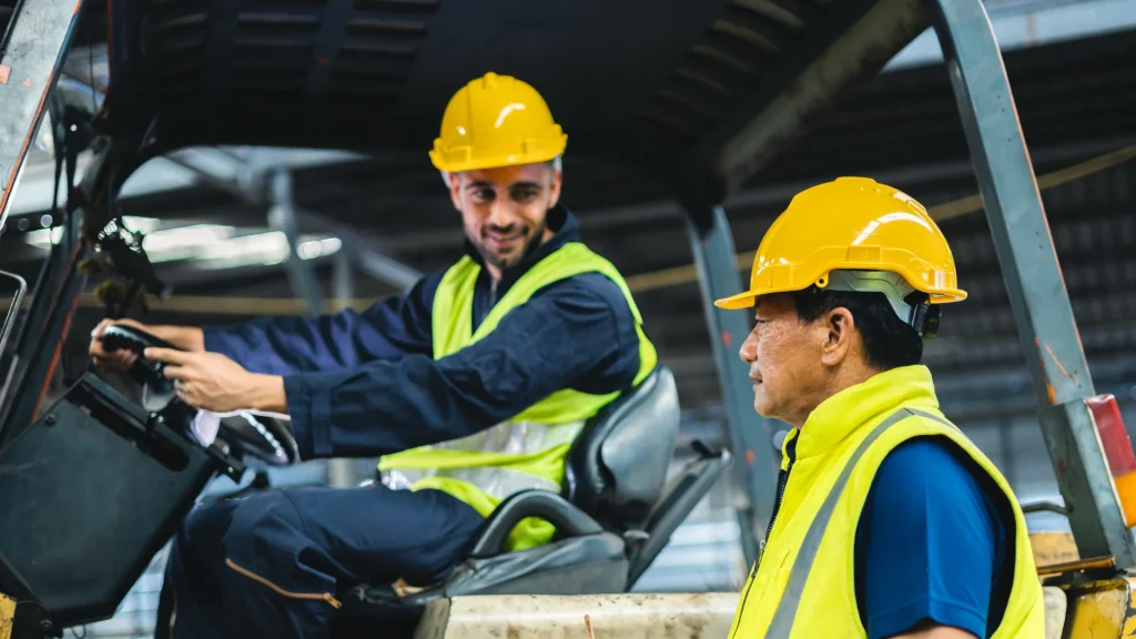 Two workers in neon safety vests and yellow helmets inside an industrial cockpit; one sits at the controls, the other stands nearby.