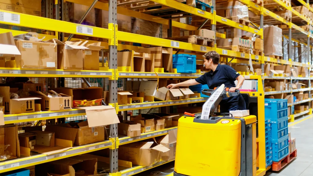 Warehouse worker operates a yellow pallet truck among tall shelves filled with boxes and packages, organizing inventory.